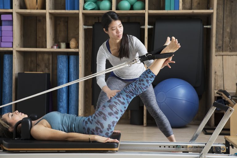Pilates teacher assists student using reformer for leg exercises.